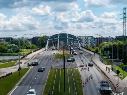 Most z ruchem drogowym, otoczony zielenią i budynkami, w tle chmury.