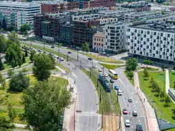 Widok na ruchliwą ulicę z tramwajem i samochodami, otoczoną nowoczesnymi budynkami i zielenią.