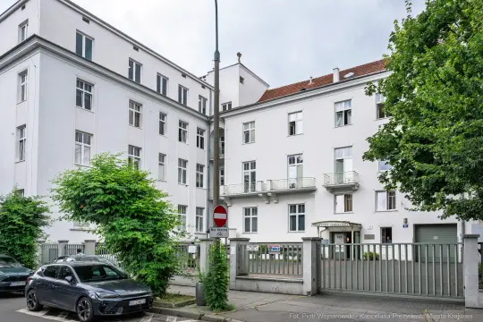 Budynek biurowy z białą elewacją, otoczony zielenią i zaparkowanymi samochodami.