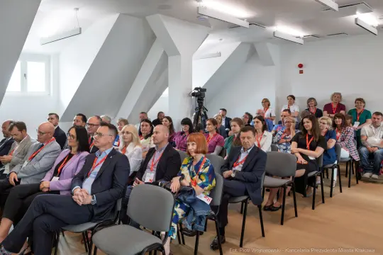 Grupa ludzi siedzących na sali konferencyjnej, uczestniczących w wydarzeniu.