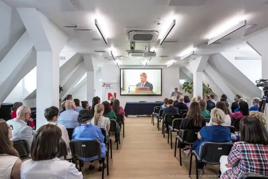 Konferencja z prelegentem na dużym ekranie, uczestnicy siedzą w sali.