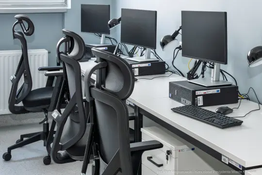 Puste stanowiska komputerowe z monitorami i ergonomicznymi krzesłami.