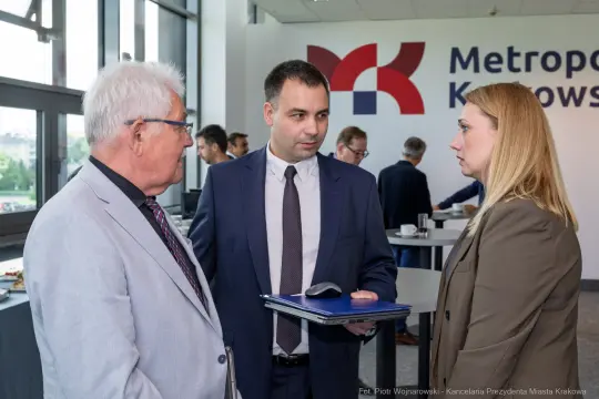 Trzech ludzi rozmawia w biurze, w tle logo Metropolii Krakowskiej.
