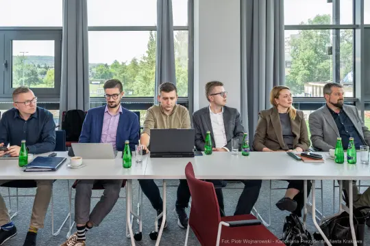 Grupa osób siedzących przy stole w sali konferencyjnej, z laptopami i butelkami wody.