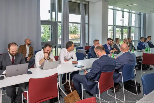 Spotkanie biznesowe w sali konferencyjnej, uczestnicy przy stołach z laptopami i notatkami.