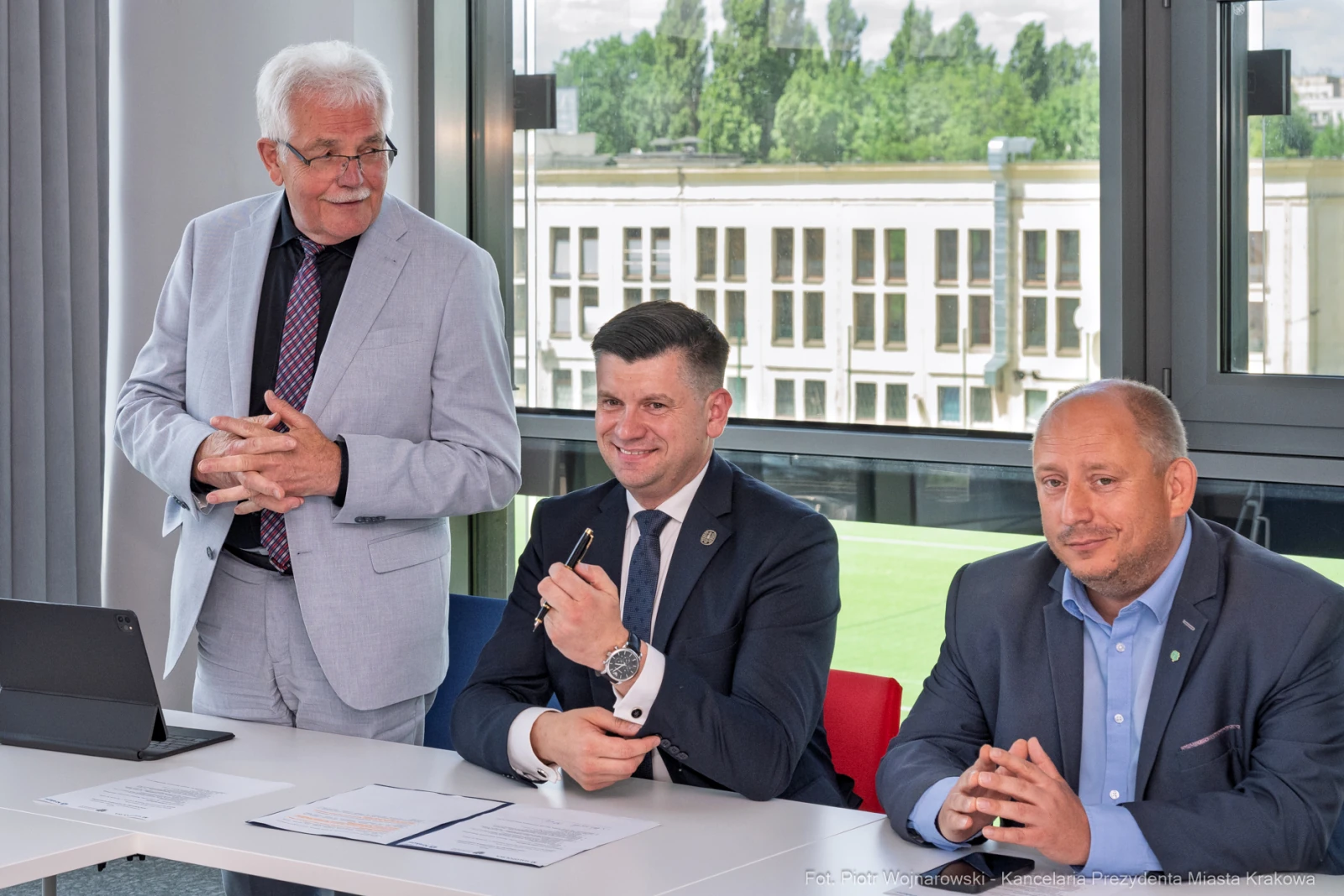 Trzech mężczyzn przy stole, jeden z nich trzyma długopis, w tle okna z widokiem na miasto.  Autor: Piotr Wojnarowski