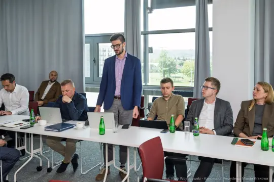 Mężczyzna w garniturze przemawia podczas spotkania w sali konferencyjnej. Obecni uczestnicy przy stole.