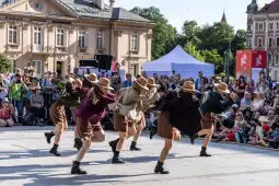 Grupa tancerzy w strojach w stylu retro wykonuje układ taneczny na ulicy, wokół tłum widzów.
