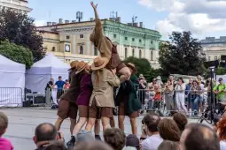 Grupa tancerzy wykonuje układ taneczny na placu, otoczona widownią.