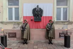 Dwaj żołnierze w mundurach stoją przed flagą Polski i pomnikiem orła.