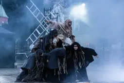 Grupa osób w kostiumach teatralnych, w otoczeniu mgły, wykonuje układ taneczny.
