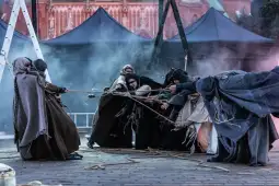 Grupa osób w strojach historycznych ciągnie linę w otoczeniu dymu i ciemnych namiotów.