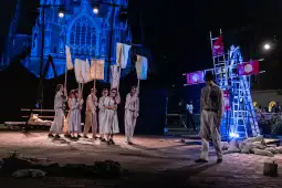 Grupa aktorów na scenie z flagami, w tle oświetlona katedra.