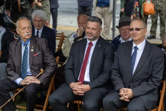 Trzech mężczyzn w garniturach siedzących na krzesłach podczas uroczystości.