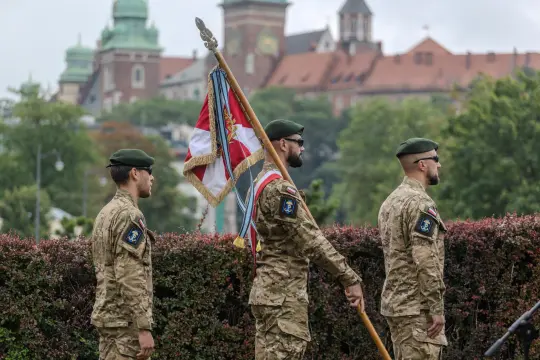 Żołnierze w mundurach wojskowych z flagą, tło z budynkami i zielenią.
