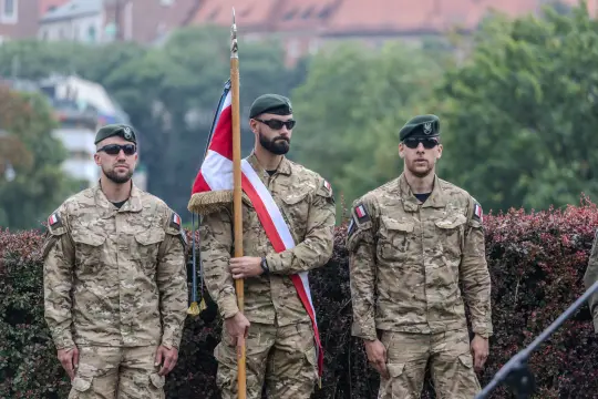 Trzej żołnierze w mundurach wojskowych, jeden z flagą, stoją w postawie defiladowej.