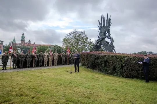 Uroczystość z udziałem żołnierzy i mowy przy pomniku w tle.