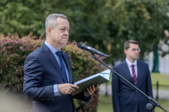 Mężczyzna w garniturze trzyma dokumenty i przemawia do mikrofonu, obok stoi drugi mężczyzna.