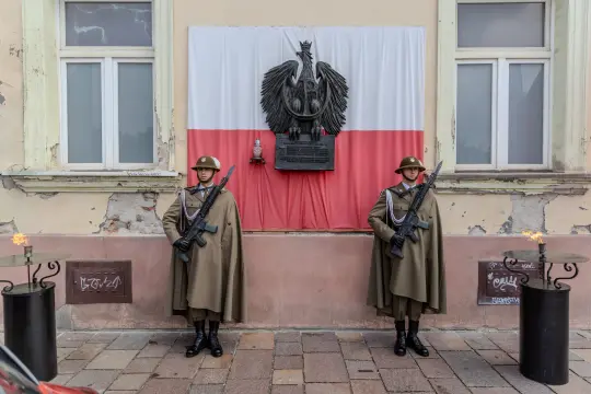 Dwaj żołnierze w mundurach stoją przed flagą Polski i pomnikiem z orłem.