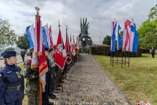 Uroczystość z flagami narodowymi i pomnikiem w tle.