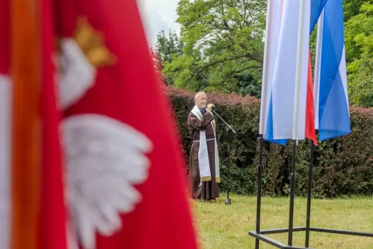 Mężczyzna w habicie przemawia przy flagach Polski i innych krajów.