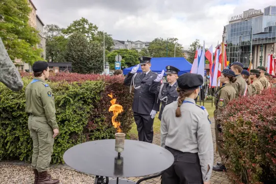 Ceremonia z udziałem strażaków, flagi narodowe i płonący znicz.