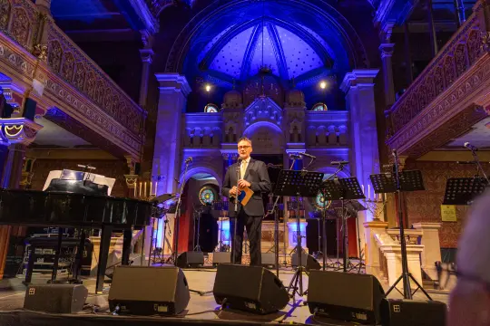 Mężczyzna z instrumentem na scenie w eleganckim wnętrzu z niebieskim oświetleniem.