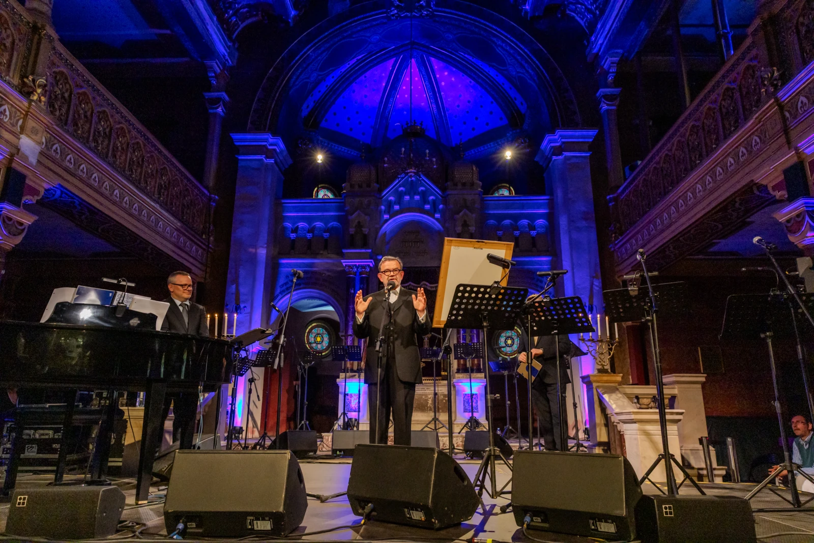 Koncert w eleganckiej sali z pianistą, mówcą i muzykami w tle. Oświetlenie w odcieniach niebieskiego.  Autor: Bogusław Świerzowski
