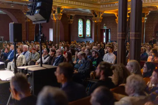 Tłum ludzi siedzących w sali, z widocznymi oknami witrażowymi i sceną.