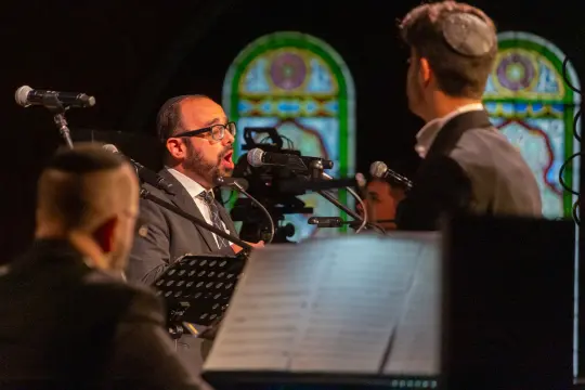 Mężczyzna śpiewa na scenie, w tle instrumenty muzyczne i kolorowe witraże.