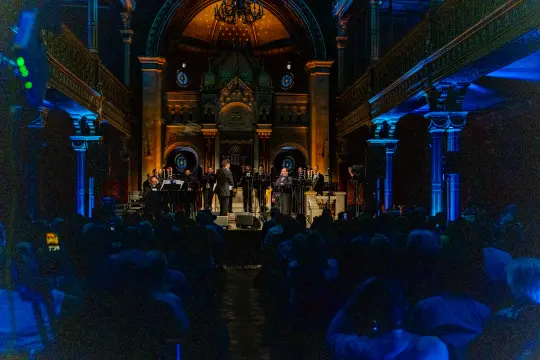 Koncert w zabytkowej sali, z chórem i orkiestrą, oświetlenie w niebieskich i pomarańczowych tonach.
