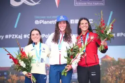 Trzy zawodniczki na podium z medalami i kwiatami, uśmiechnięte, w strojach reprezentacyjnych.