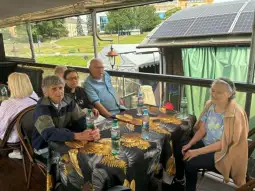 Grupa osób siedzących przy stole na tarasie, w tle widoczna zieleń i budynki.