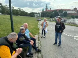 Grupa osób rozmawia na chodniku w pobliżu rzeki, w tle zamek i zielone wzgórza.