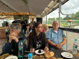 Grupa osób siedzących przy stole na pokładzie łodzi, jedzących ciasto i rozmawiających.
