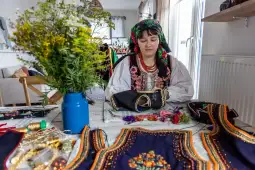 Kobieta w tradycyjnym stroju szyje hafty, wokół niej materiały i kwiaty w wazonie.