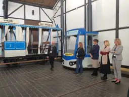 Mężczyzna prowadzi grupę kobiet obok historycznego tramwaju i nowoczesnego pojazdu.