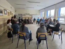 Grupa ludzi siedzących w kręgu w jasnej sali.