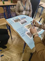 Osoba pisząca na niebieskim plakacie z zdjęciami i notatkami, w tle uczestnicy spotkania.