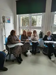 Cztery kobiety w sali szkoleniowej, siedzące przy biurkach, rozmawiają ze sobą.