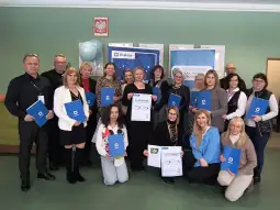 Grupa ludzi trzymająca dyplomy i certyfikaty w sali z dekoracjami.