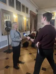 Kobieta przemawia do dziennikarza w sali z portretami na ścianach.