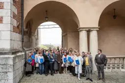 Grupa ludzi pozująca do zdjęcia, trzymająca niebieskie teczki, w architektonicznym otoczeniu.