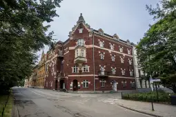 Czerwony budynek z detalami architektonicznymi na rogu ulicy, otoczony drzewami.