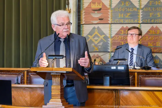 Mężczyzna przemawia z mównicy w sali obrad, obok siedzi inny mężczyzna.