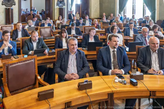Spotkanie w sali obrad z uczestnikami przy stołach, z laptopami i dokumentami.