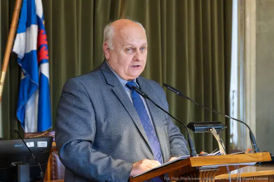 Mężczyzna przemawia przy mównicy, w tle flagi i sprzęt multimedialny.