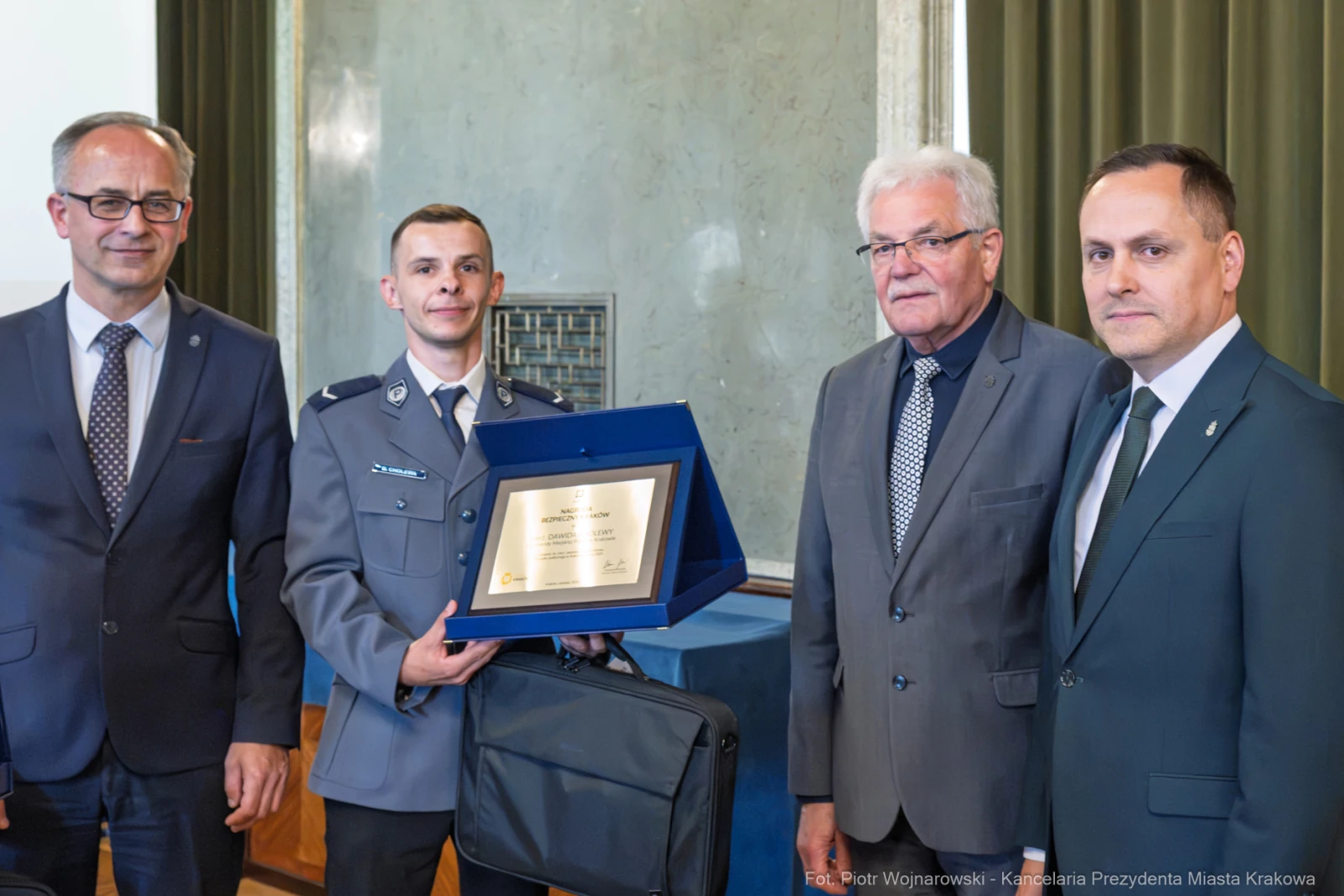 Czterech mężczyzn w garniturach, jeden z odznaczeniem w ręku, w tle ściana z zielonymi zasłonami.  Autor: Piotr Wojnarowski