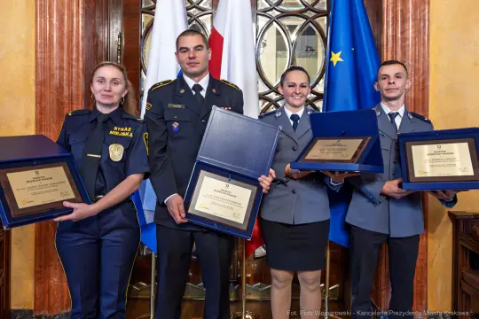 Cztery osoby w mundurach trzymają dyplomy i nagrody w eleganckim wnętrzu.