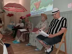 Cztery osoby w kapeluszach siedzą na krzesłach, czytając teksty, w tle parasole i dekoracje.
